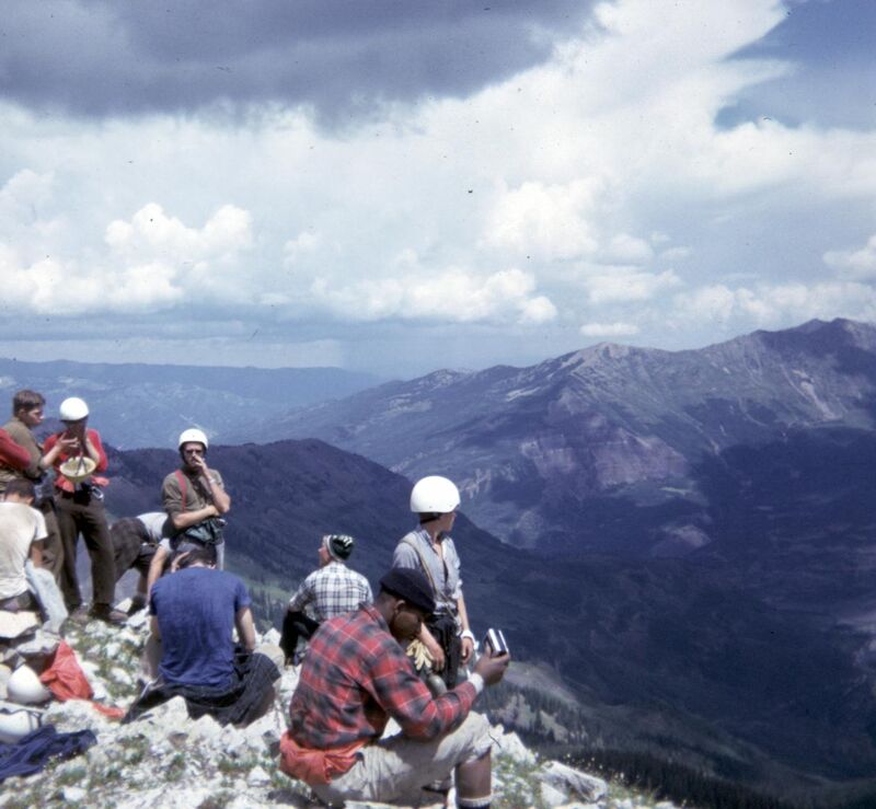 A group of people are resting on a rocky mountain peak, enjoying the scenic view of the surrounding mountains and valleys under a cloudy sky. Some are sitting, while others are standing, and they appear to be taking a break during a hike or climb. The atmosphere suggests a moment of camaraderie and appreciation for nature's beauty.